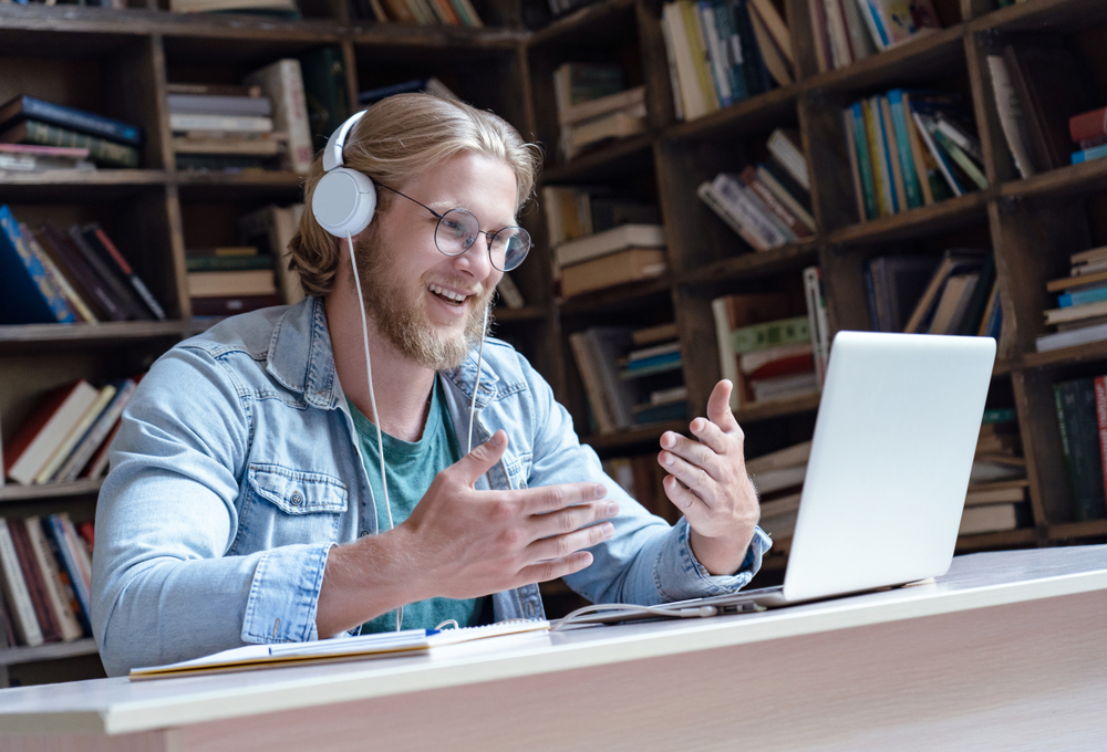 Happy male student online teacher wear headphone talk video calling looking at laptop computer screen do conference chat communicate with skype tutor, distance education e learning course in library