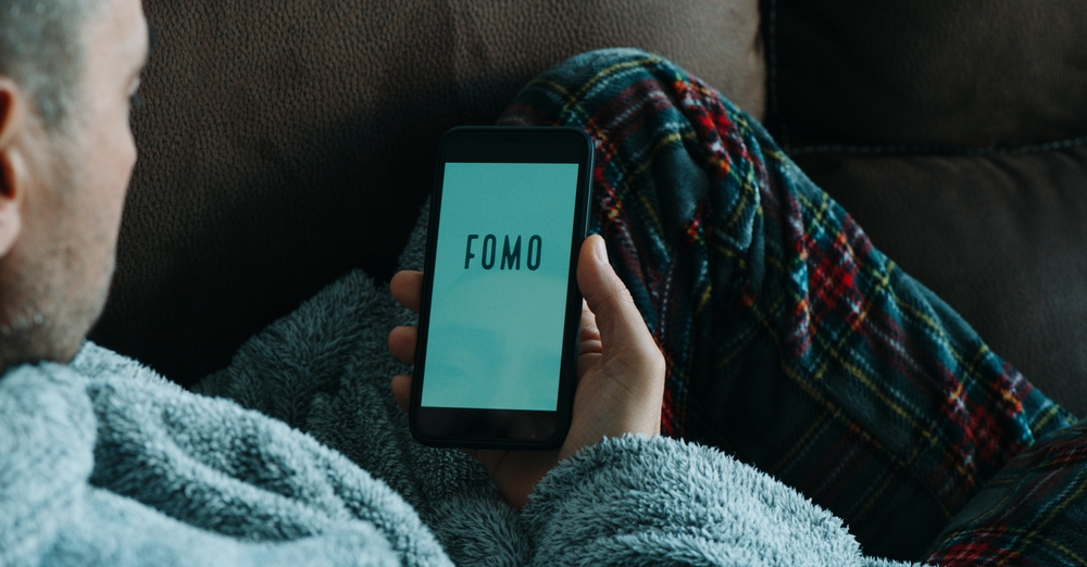 a caucasian man, lying in the sofa wearing pajamas and house robe, reads the text fomo, standing for fear of missing out, in his smartphone, in a panoramic format to use as web banner or header
