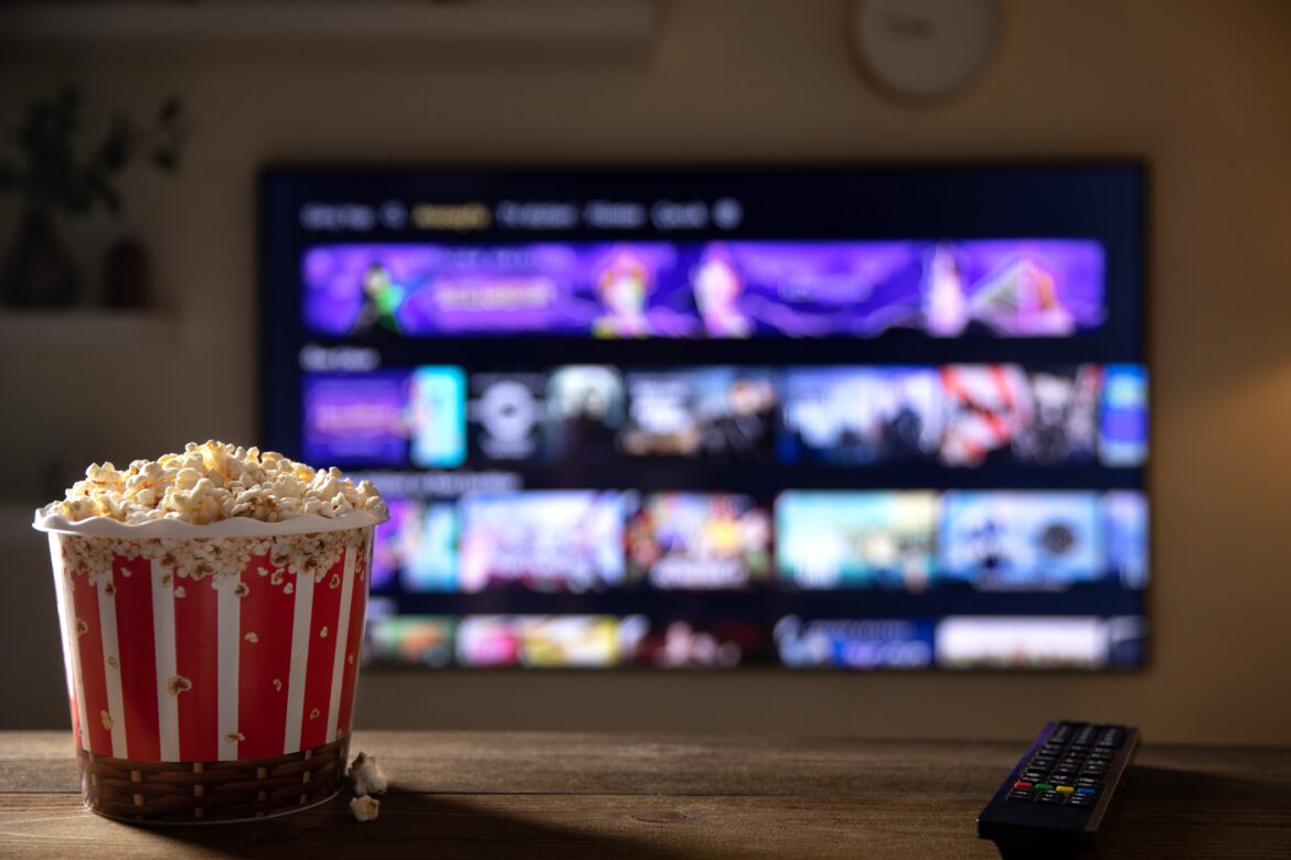 Close up of bowl of popcorn and remote control with tv works on background. Evening cozy watching a movie or TV series at home.