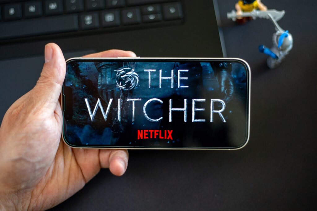 Hand holding a TV remote with The Witcher logo on Netflix, surrounded by playful miniatures and a laptop keyboard. Addicted to this captivating TV series based on the popular game franchise.