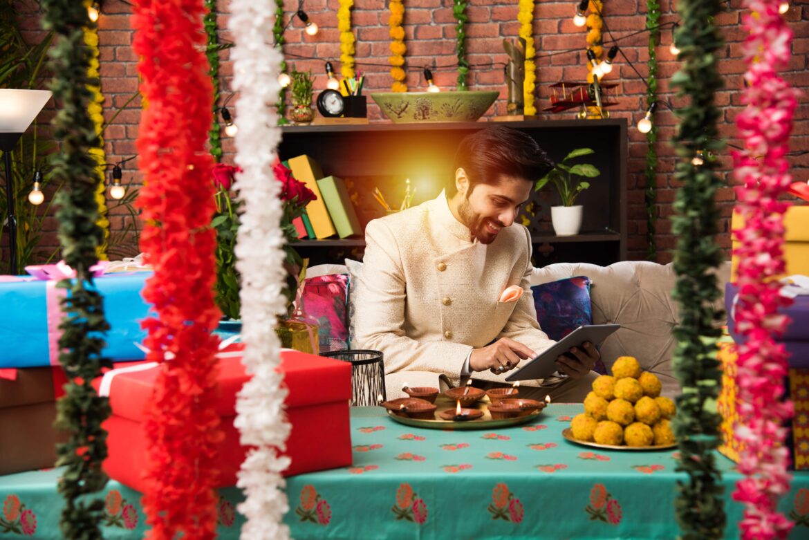 Indian man using tablet touchscreen computer on festival day while wearing traditional outfit. sitting on sofa with gifts and sweets with lighting bokeh. Online shopping of gadgets on Diwali sale
