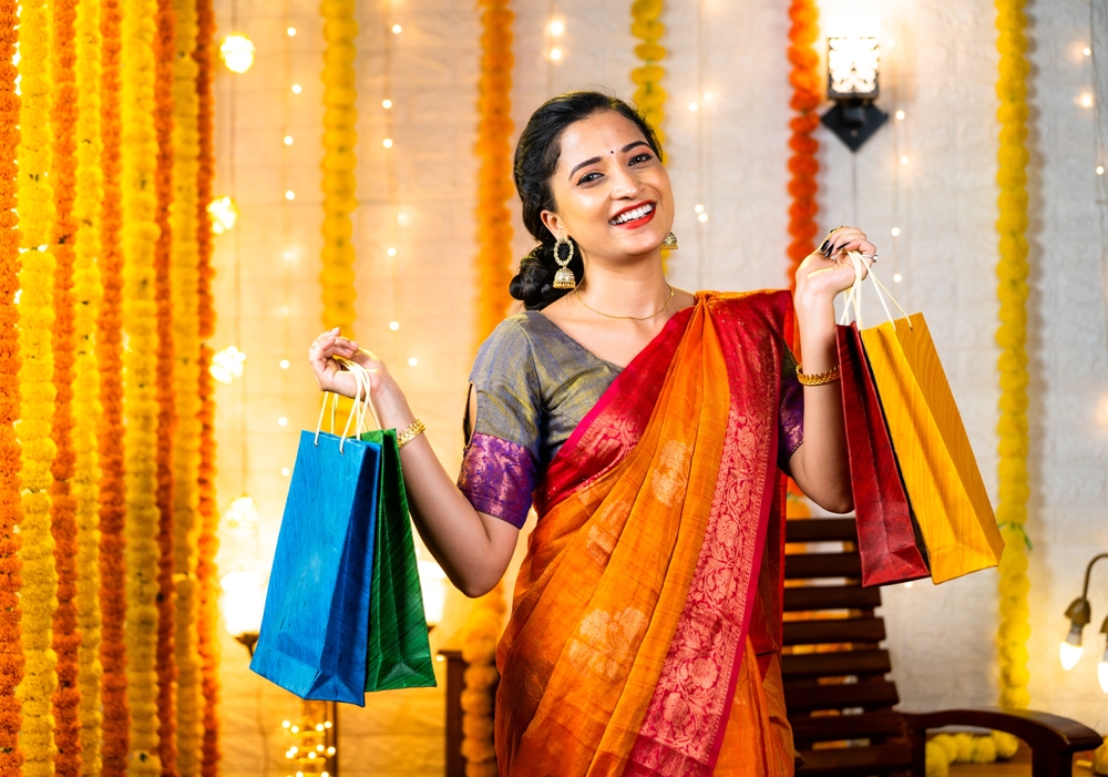 Happy young indian girl in saree with shopping bags in hand looking at camera at home - concept of festival offers, promotional sales and discount