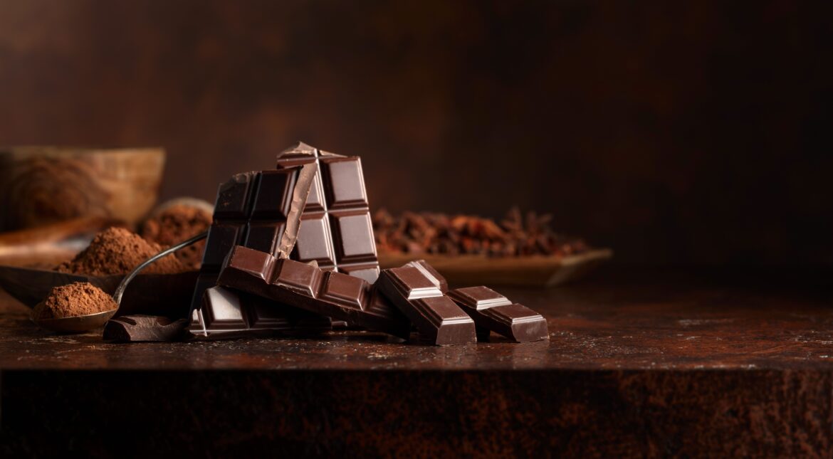 Broken dark chocolate bar and cocoa powder on a brown table. Copy space.